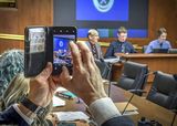 Representative Lillie holds up his smartphone to take a photo of Representative Fischer, who is sitting at the testifiers' table.
